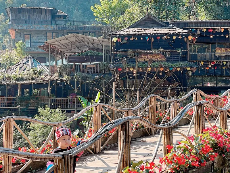 A toddler in a traditional hat stands on a wooden bridge overlooking waterwheels and cafes in Cat Cat Village. A toddler in a traditional hat stands on a wooden bridge overlooking waterwheels and cafes in Cat Cat Village.