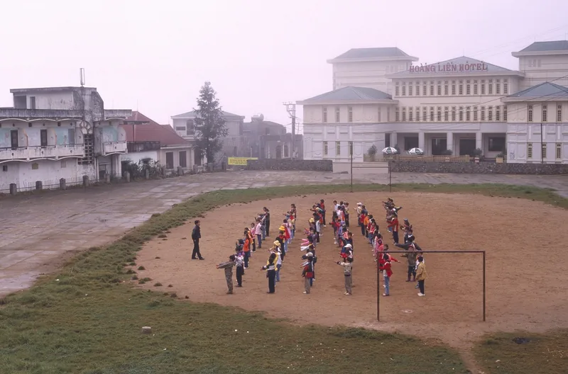 Children line up for morning exercises on a dirt square in Sapa, with Hoang Lien Hotel in the misty background. Children line up for morning exercises on a dirt square in Sapa, with Hoang Lien Hotel in the misty background.