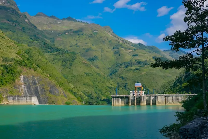 A hydroelectric dam spans a turquoise reservoir surrounded by green mountains along the Ha Giang Loop.