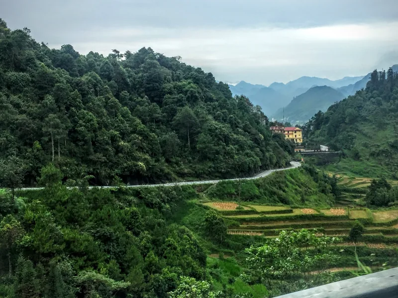 A winding mountain road curves through forested hills and terraced fields near Tam Son, Ha Giang.