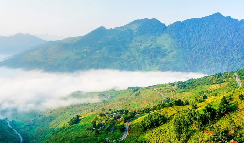 A winding mountain road cuts through green valleys and mist-covered peaks in Ha Giang.