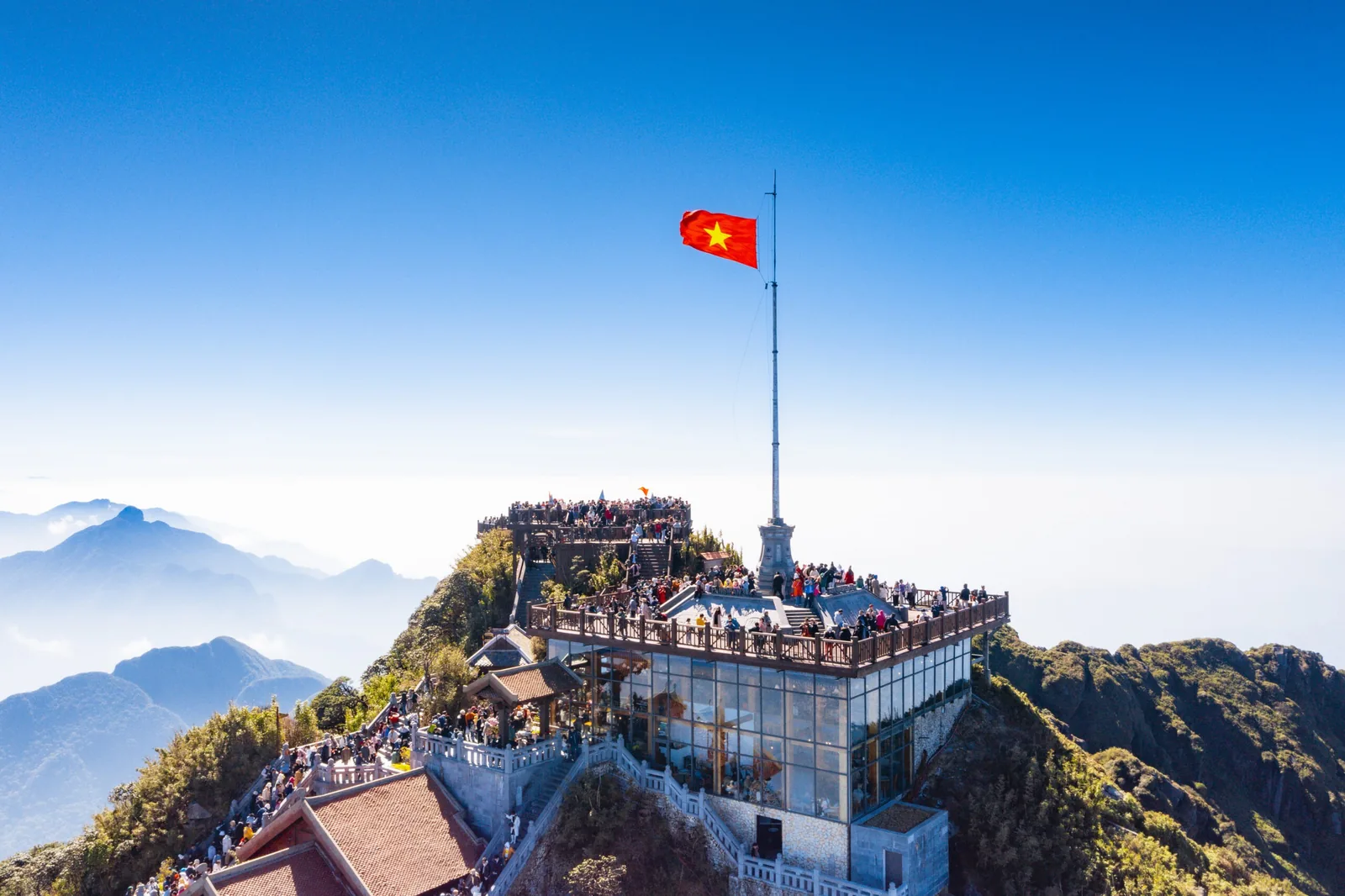 Crowds gather beneath the Vietnamese flag atop Fansipan peak, with mountain ridges rising through the clouds below. Crowds gather beneath the Vietnamese flag atop Fansipan peak, with mountain ridges rising through the clouds below.