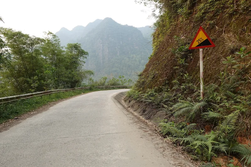 A winding mountain road with a steep gradient warning sign curves past limestone peaks near Pu Luong.