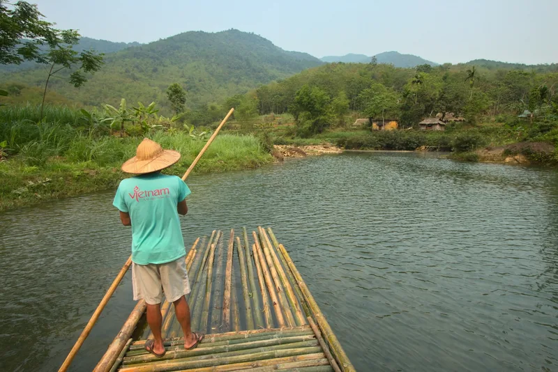 A man in a conical hat poles a bamboo raft across a calm river surrounded by hills at Pu Luong. A man in a conical hat poles a bamboo raft across a calm river surrounded by hills at Pu Luong.