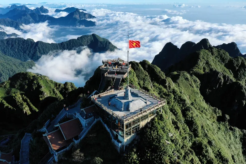 A Vietnamese flag flies atop the Fansipan summit station, surrounded by jagged peaks and a sea of clouds.