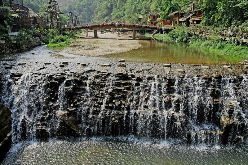 A stone waterfall flows through Cat Cat Village, with a wooden bridge and traditional houses among green hills.