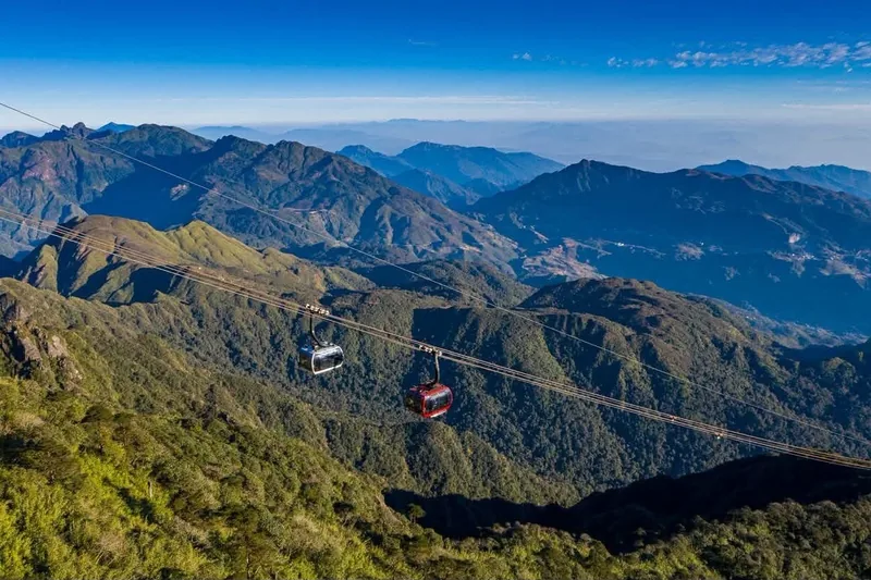 Cable car cabins glide over green mountain ridges toward Fansipan peak under a clear blue sky. Cable car cabins glide over green mountain ridges toward Fansipan peak under a clear blue sky.