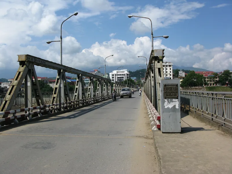 A steel truss bridge with old lampposts crosses a river leading into Sapa town under a partly cloudy sky. A steel truss bridge with old lampposts crosses a river leading into Sapa town under a partly cloudy sky.